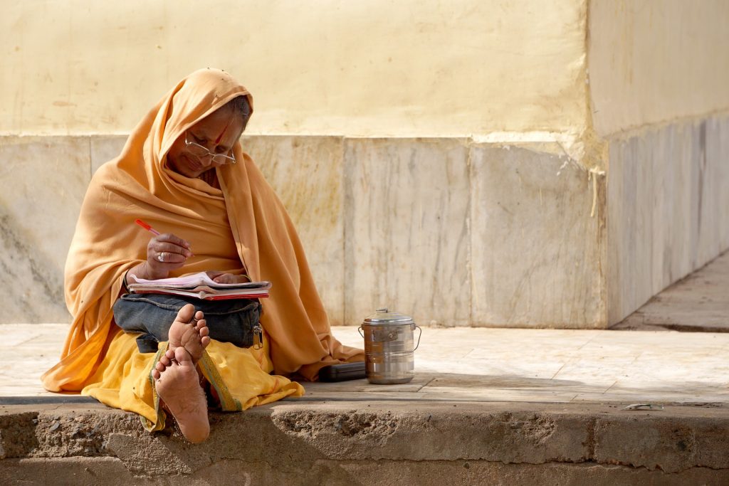 indian woman relaxed sitting on street writing.  Bare feet exposed, wearing yellow and smiling.